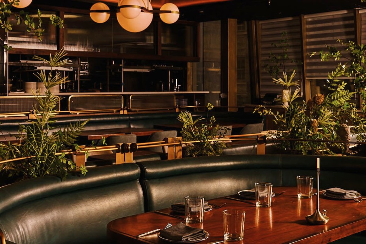 Dining room with green leather booths, wooden table set with glasses and napkins, open kitchen in background, greenery decor