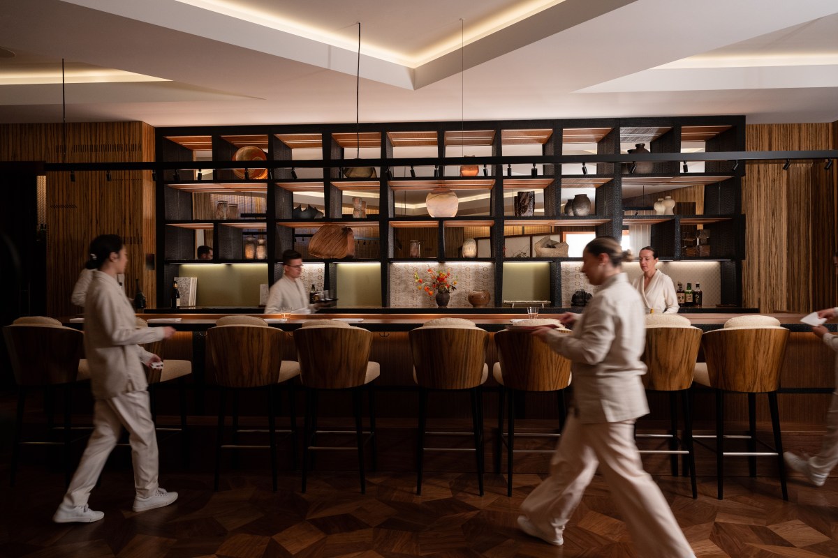Restaurant bar area with wooden stools, shelving of ceramics, and staff in white uniforms walking by