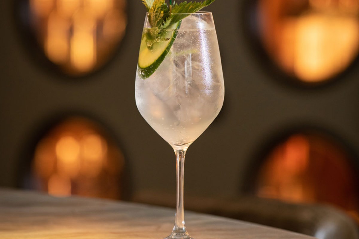 Gin and tonic cocktail with cucumber and mint garnish on a table, warm lounge lighting behind