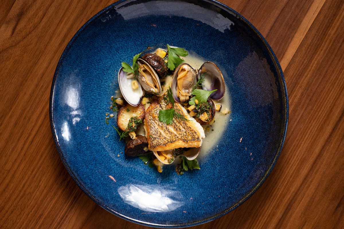 Plated seared fish with clams, corn, and herbs on a blue ceramic plate on wood table