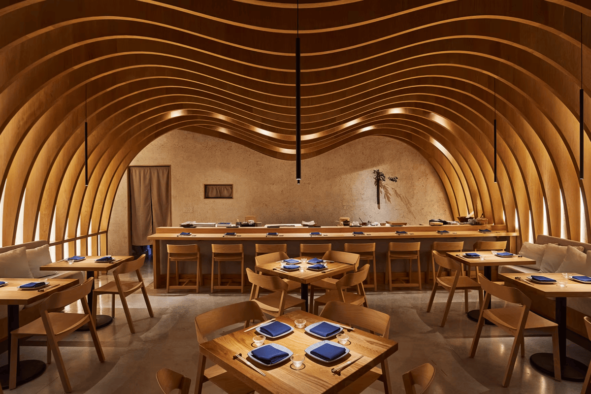 Dining room with wavy wood-ribbed ceiling, sushi counter and tables set with blue napkins
