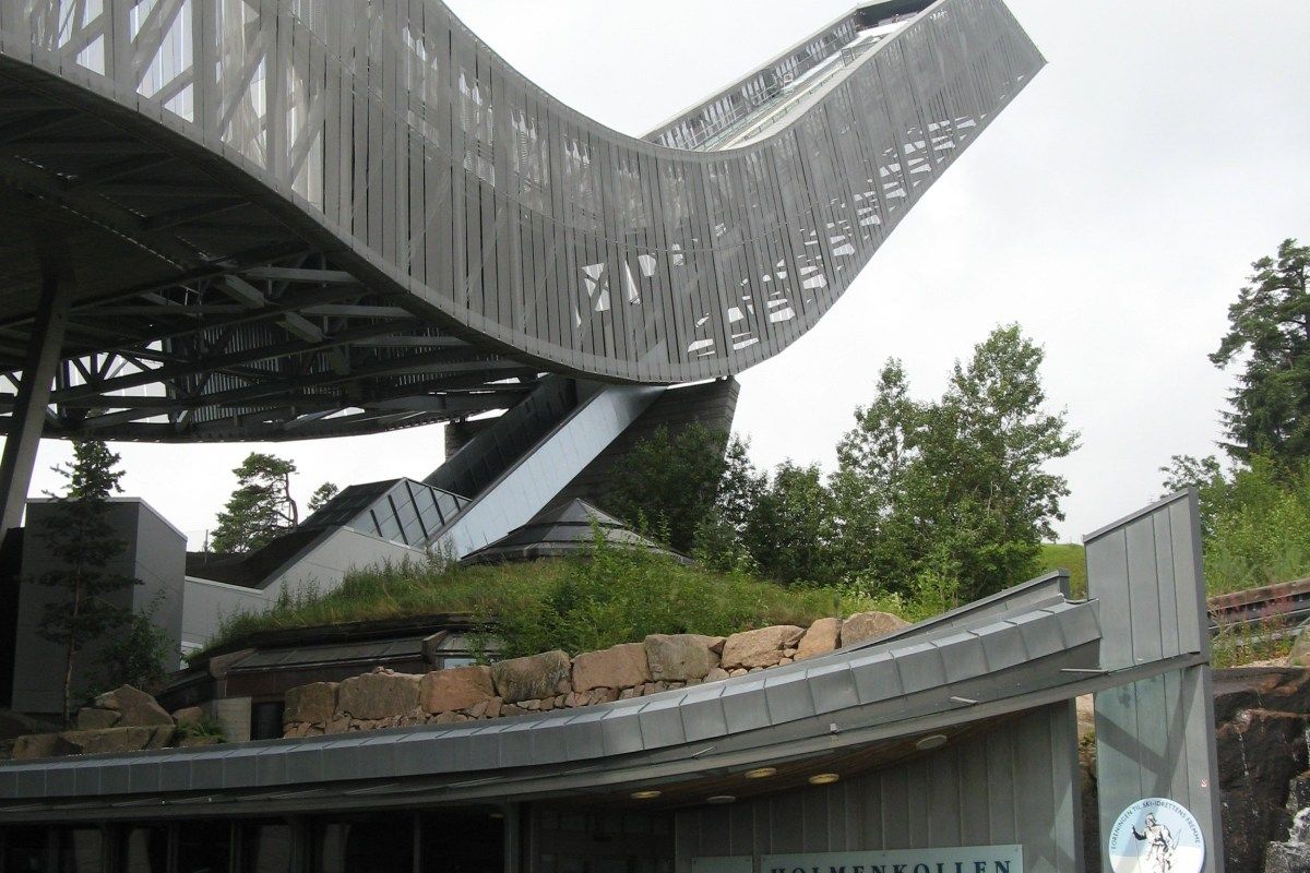 Low-angle view of the curved Holmenkollen ski jump tower with museum entrance below, green roof and stone wall.