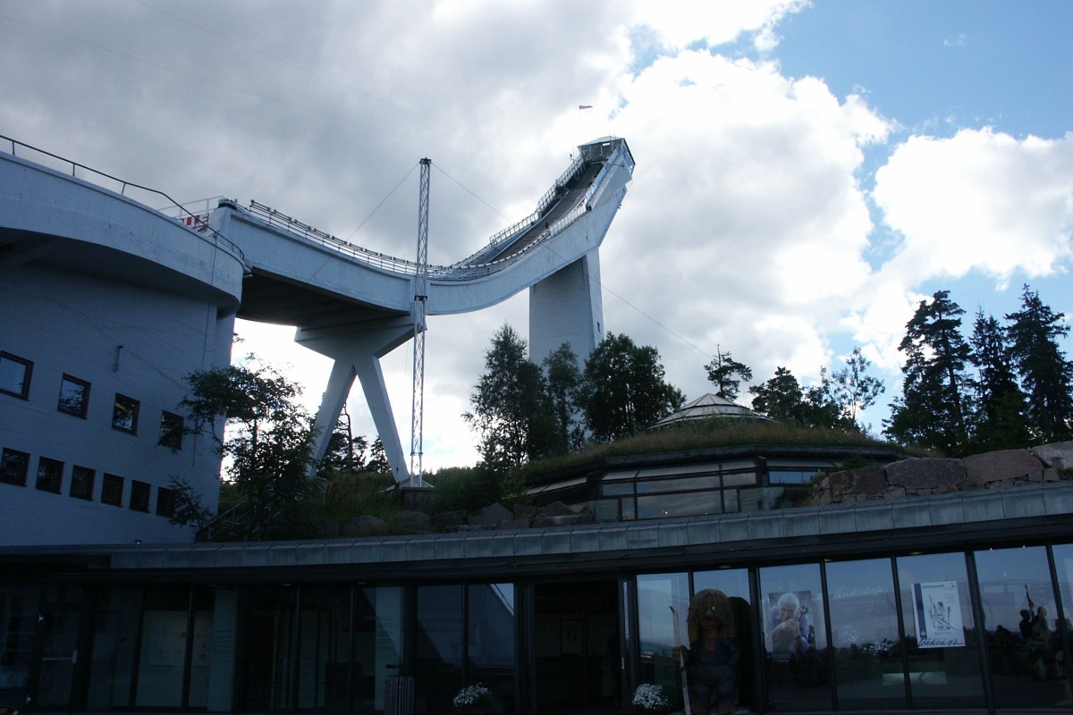 Holmenkollen ski jump tower and museum building seen against a cloudy sky, with glass dome and entrance visible.