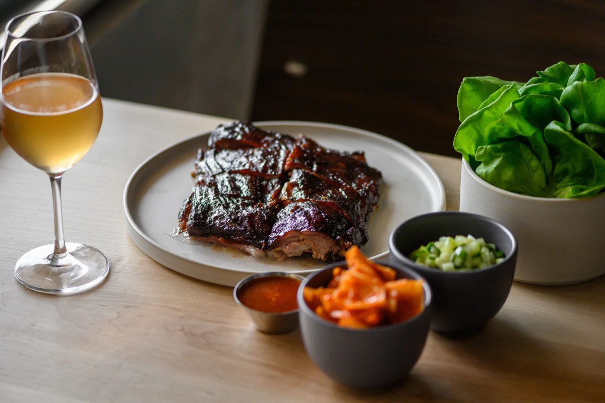 Plated barbecue ribs with dipping sauces, pickled vegetables, greens and a glass of wine on a wood table