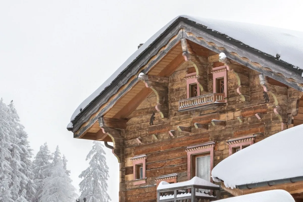 Snow-covered wooden chalet facade with pink-trimmed windows and balcony, traditional alpine architecture detail.