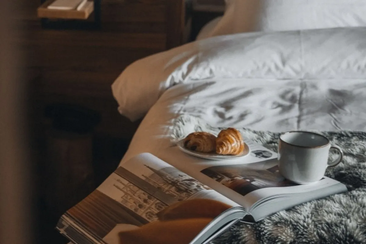 Breakfast tray with croissants, coffee cup, and magazine on a hotel bed.