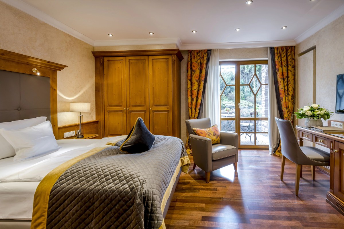 Spacious hotel guest room with large bed, wooden wardrobe, armchair, desk, and balcony door to garden