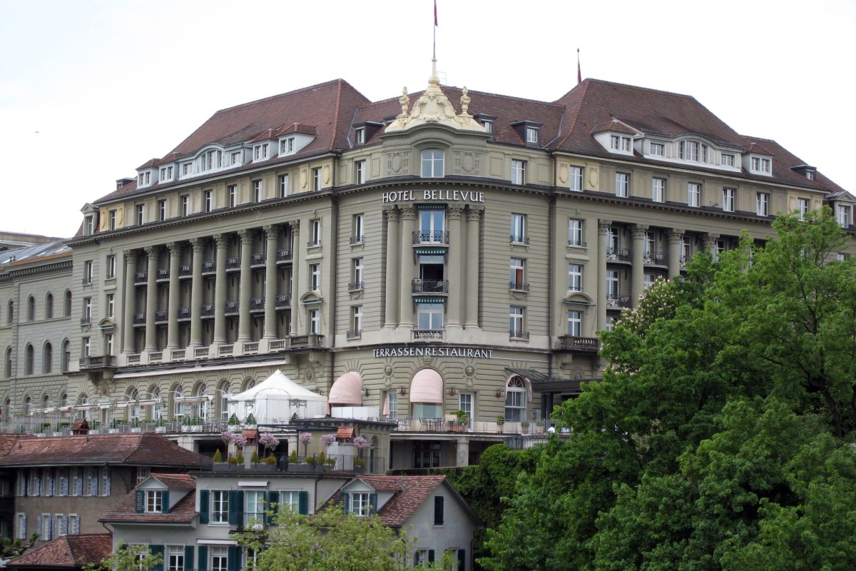 Full exterior view of Hotel Bellevue Palace facade with red-tiled roofs, terrace restaurant and surrounding greenery in Bern