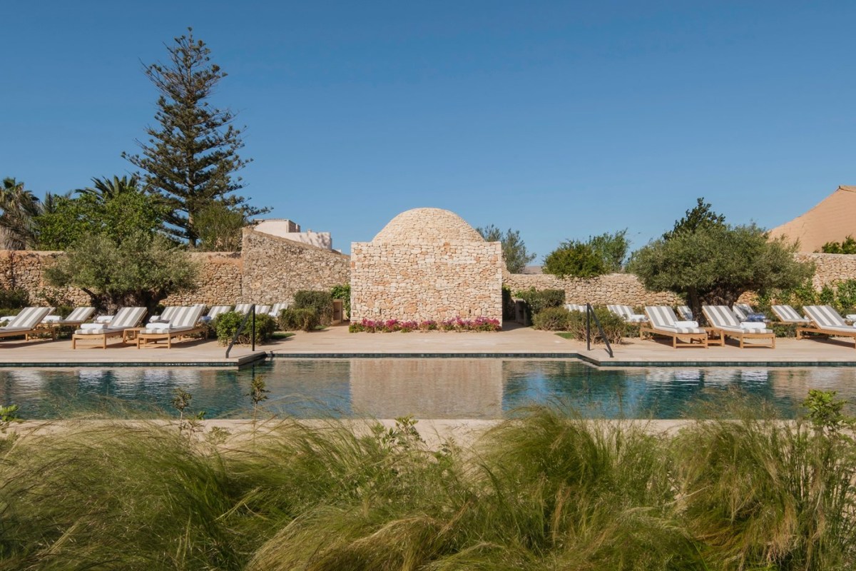Stone hotel building with domed structure beside an infinity pool and lounge chairs under blue sky
