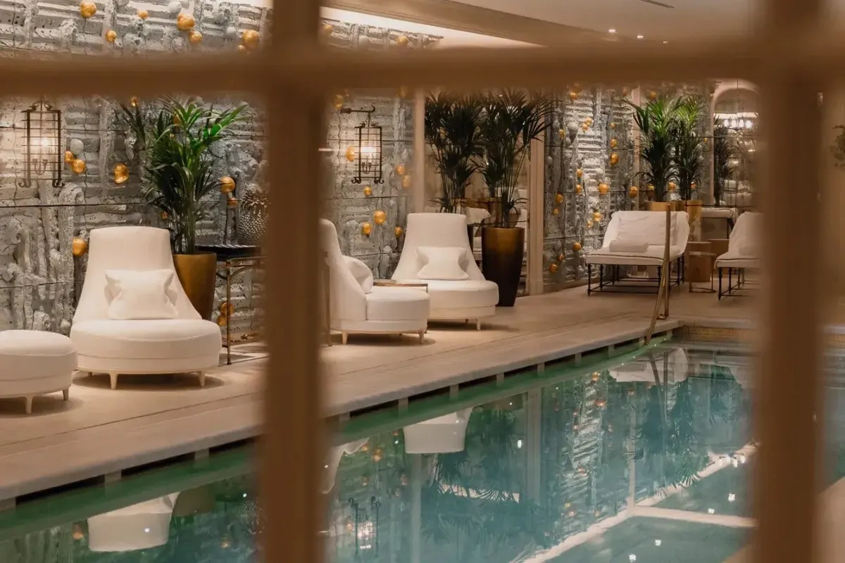 Indoor hotel pool with white lounge chairs and palm plants, viewed through a window grid