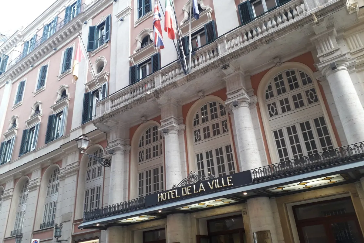 Low-angle view of the Hotel de la Ville façade in Rome, showing ornate windows, columns, flags, and the hotel's entrance sign.