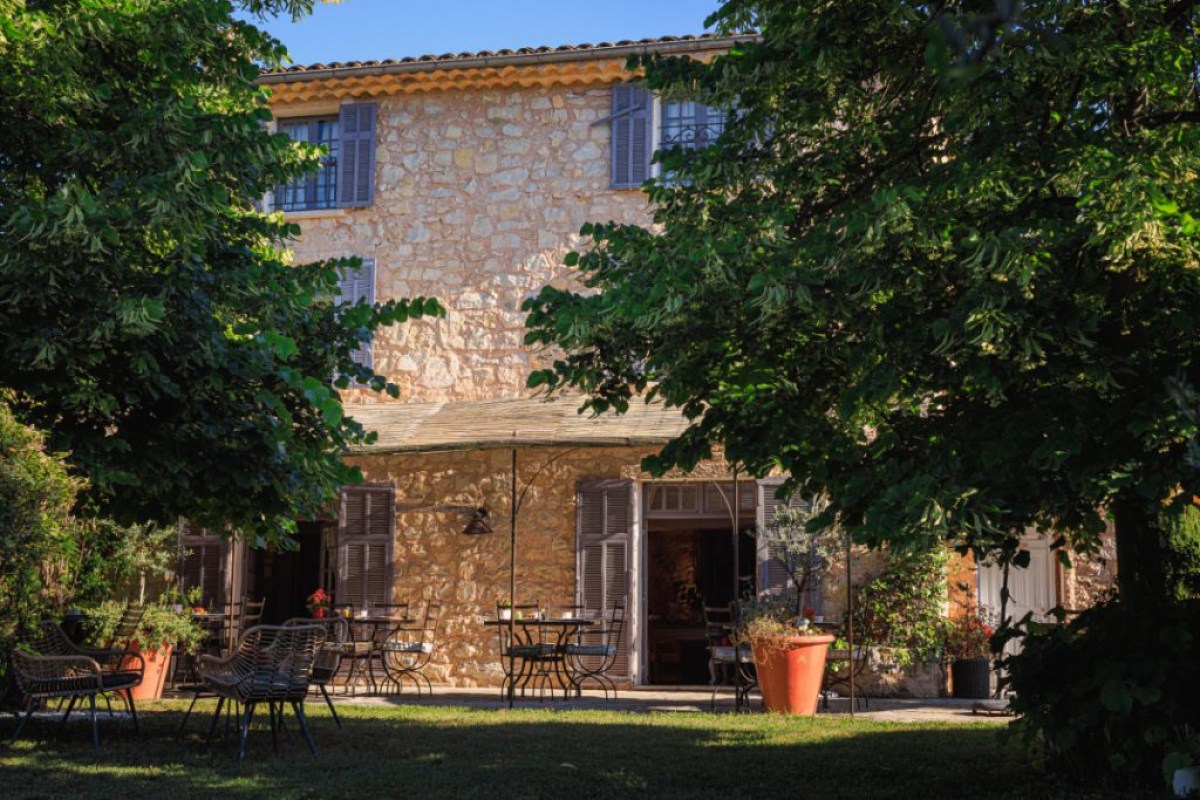 Stone façade of the hotel with shuttered windows, surrounded by trees and garden terrace with tables