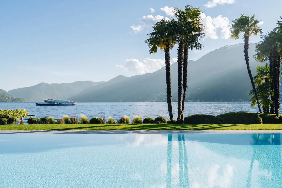 Infinity pool with palm trees overlooking Lake Maggiore and mountains, with a boat passing by