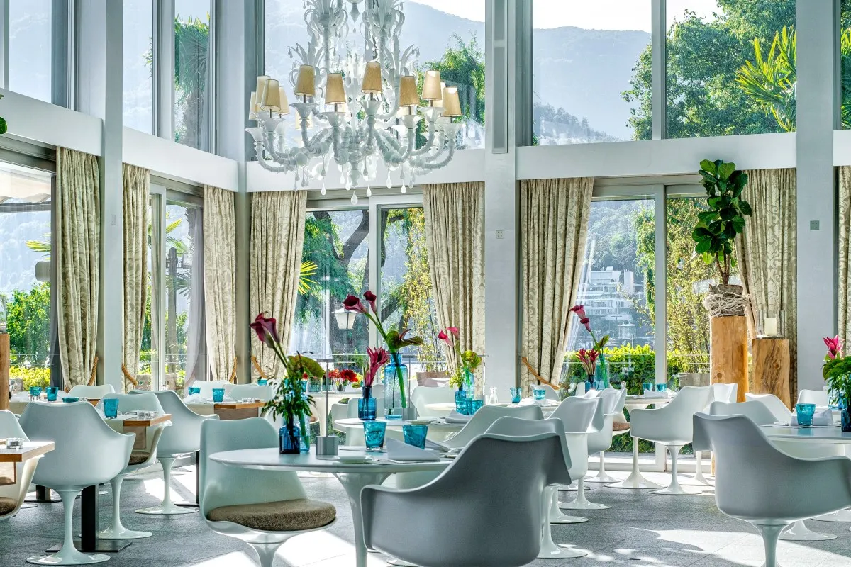 Bright hotel restaurant interior with modern white chairs, chandelier, and large windows overlooking greenery
