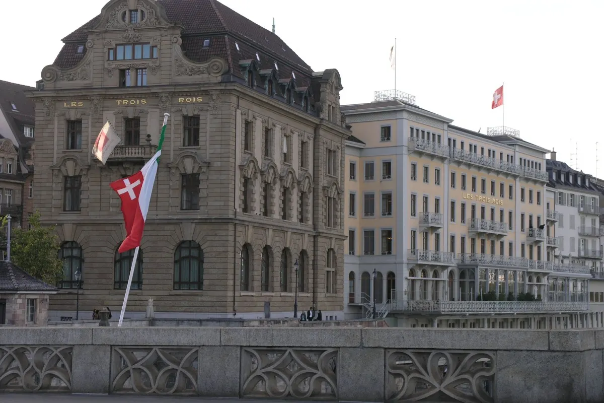 Hotel Les Trois Rois façade seen from a bridge, with Swiss and Basel flags flying above the entrance.