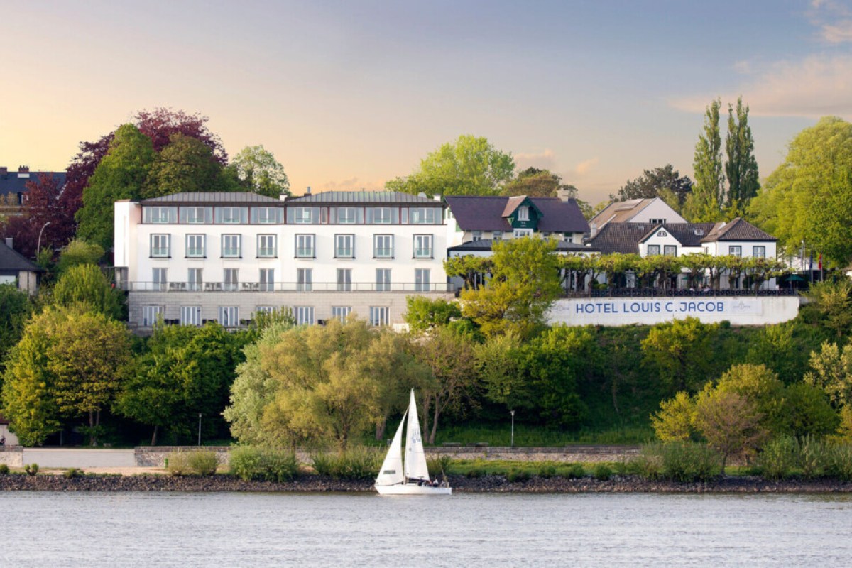 Hotel Louis C. Jacob seen from the river at sunset, with a sailboat passing by in front of the tree-lined building