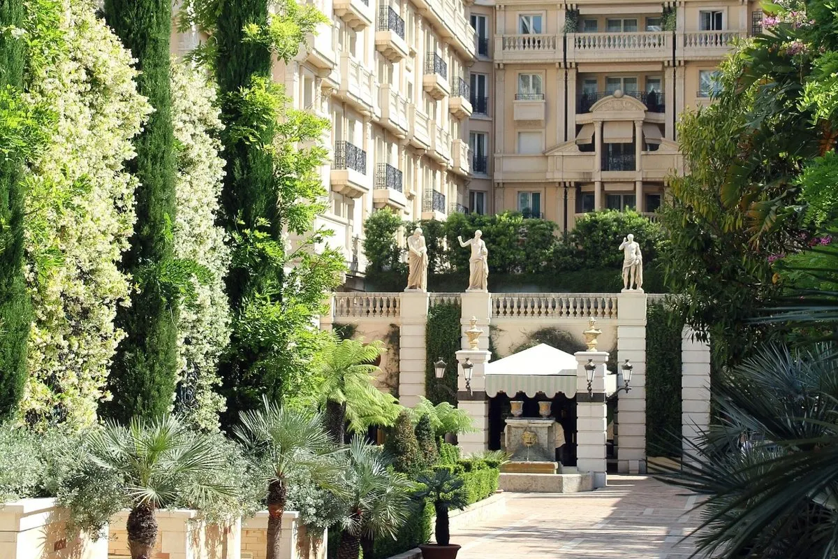 The facade and garden walkway of Hôtel Métropole Monte-Carlo, lined with cypresses and palm trees