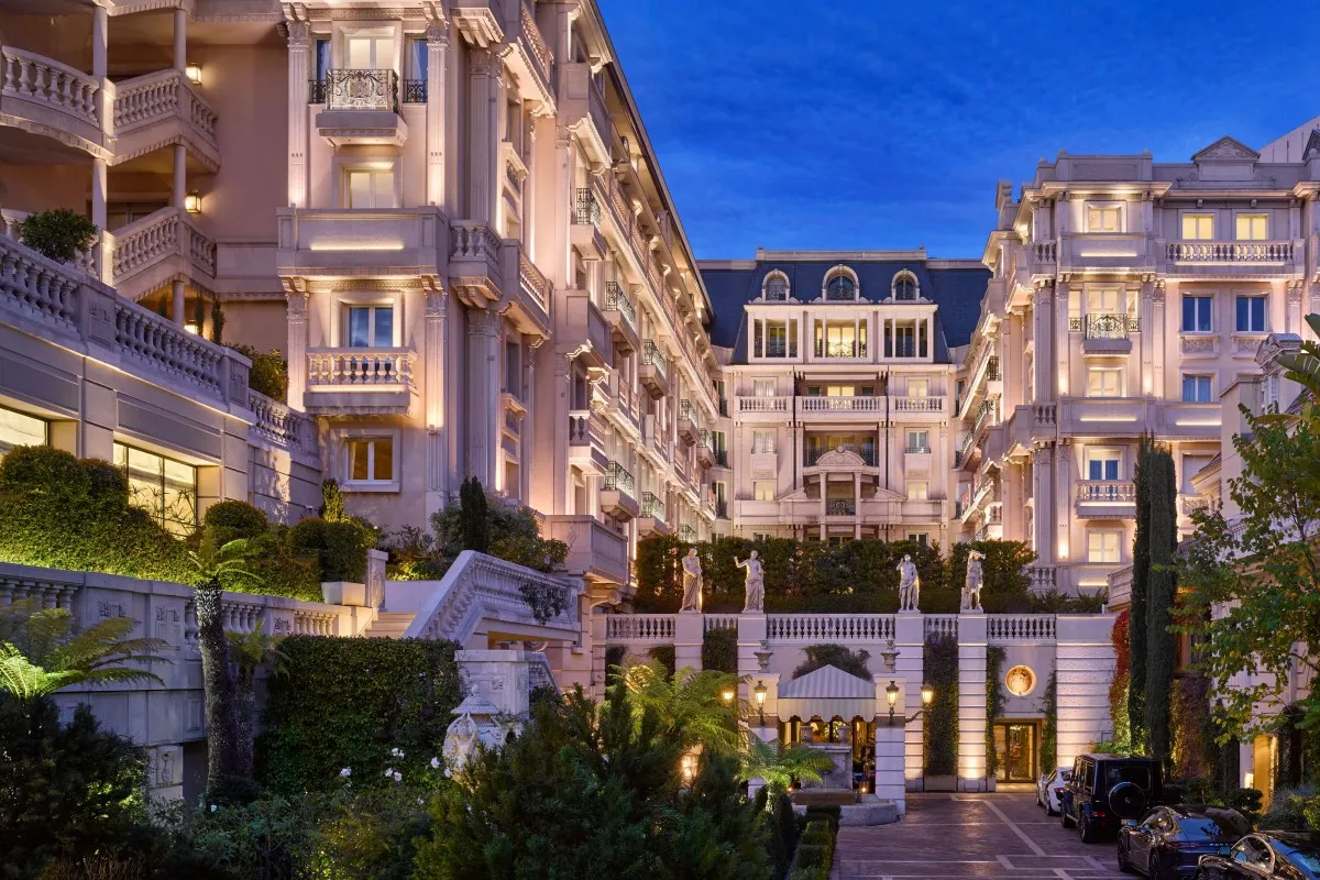 Illuminated façade of Hotel Metropole Monte Carlo at dusk, ornate balconies and entrance with gardens.