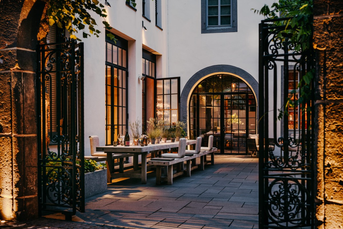 Hotel courtyard at dusk with long outdoor dining table, wrought-iron gate and illuminated building facade