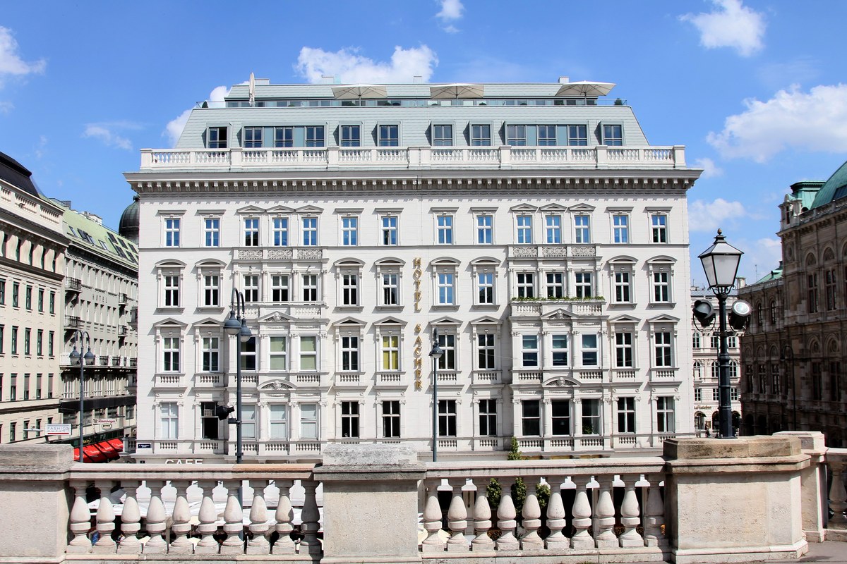 Full facade of Hotel Sacher viewed from Albertina terrace, white building under blue sky with rooftop terrace