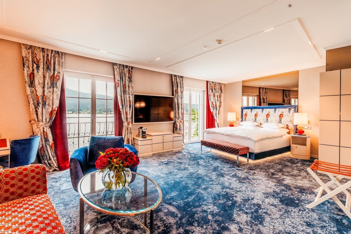 Elegant hotel suite bedroom with blue carpet, patterned bird curtains, bed, and lake-view balcony doors