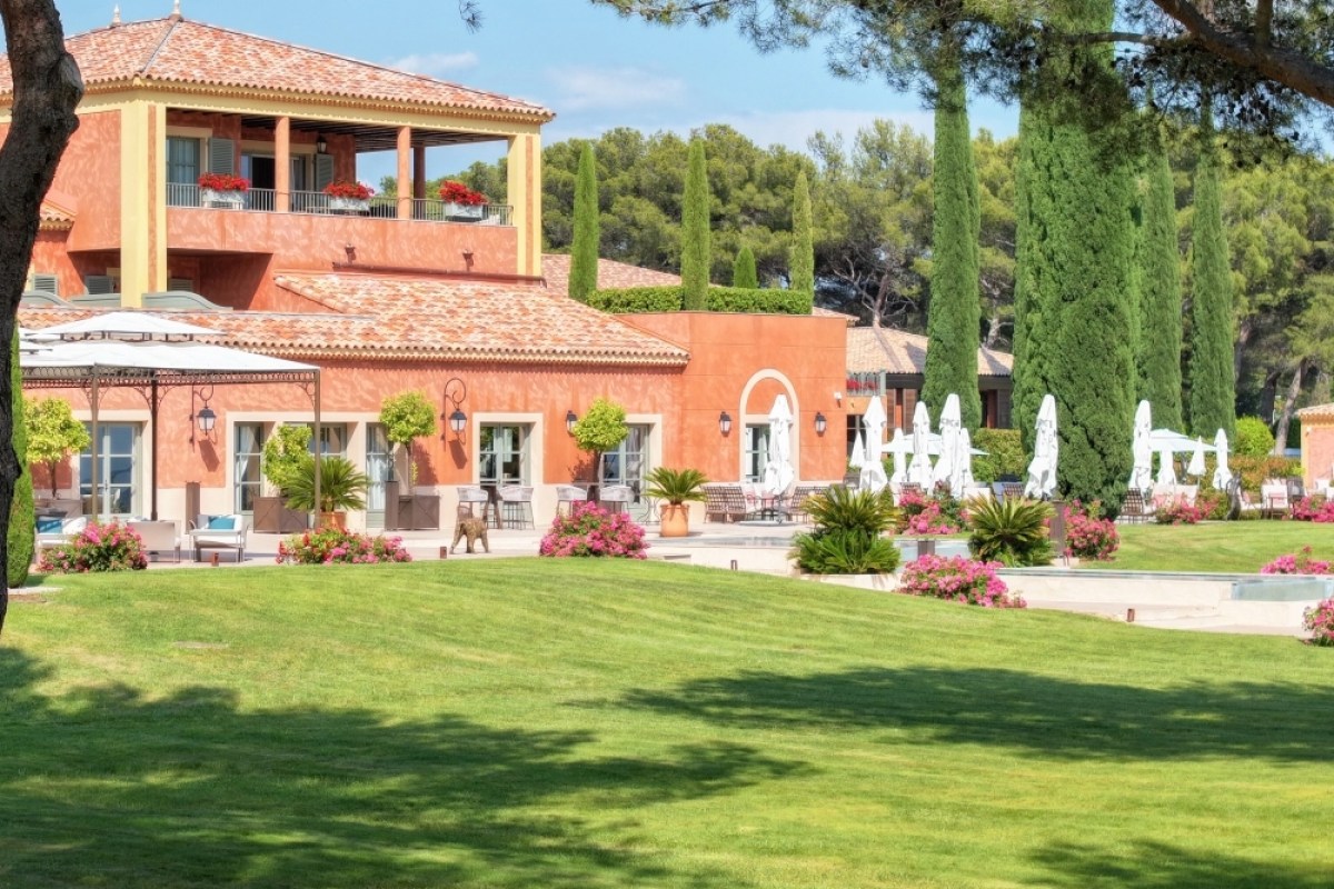 Ochre-colored hotel façade with terrace, balconies, cypress trees, lawn and pool umbrellas in a landscaped garden.