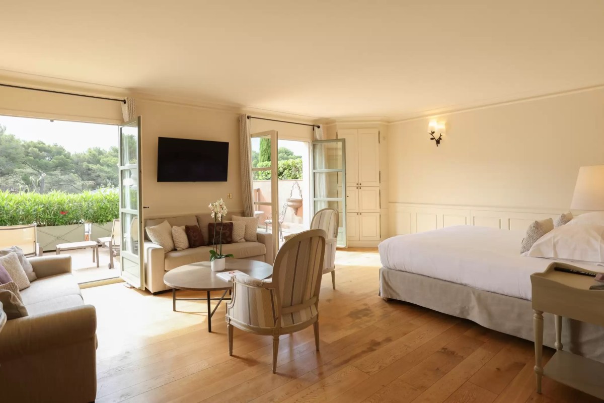 Spacious hotel suite with bed, seating area and open doors to a terrace with greenery