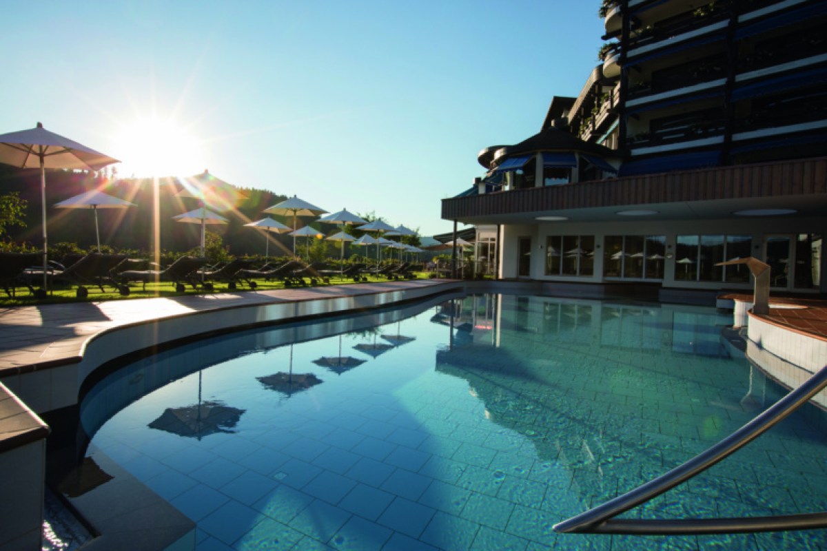 Hotel exterior with outdoor pool, loungers and umbrellas at sunset, green hills in background