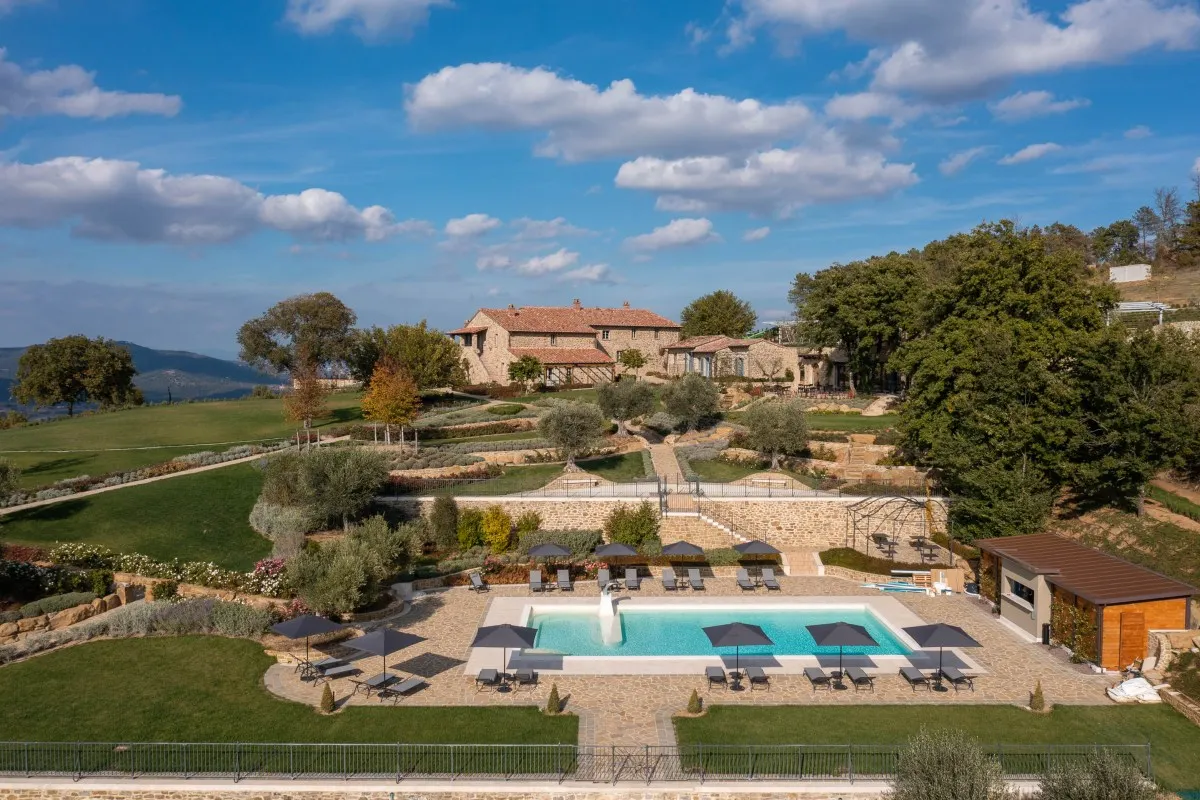 Aerial view of stone hotel buildings overlooking a large swimming pool with loungers, terraced gardens and hills
