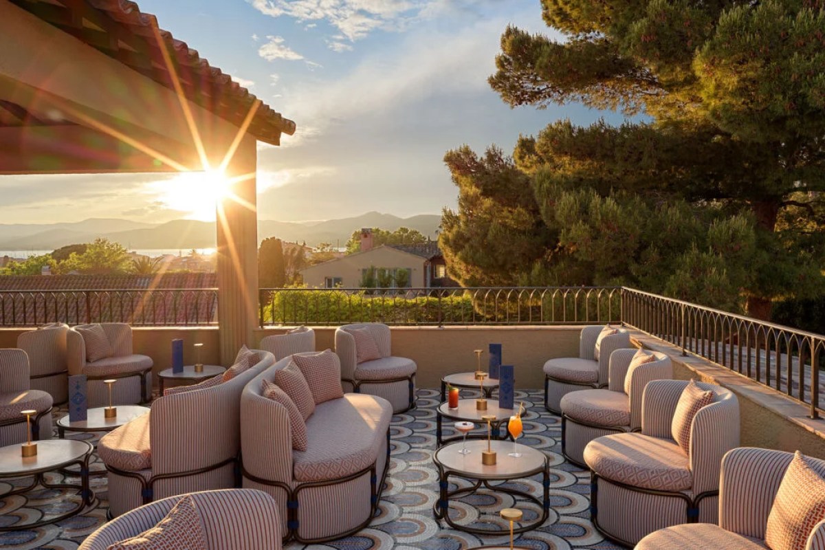 Rooftop lounge terrace at sunset with patterned sofas, round tables, cocktails and sea/mountain view through pine trees