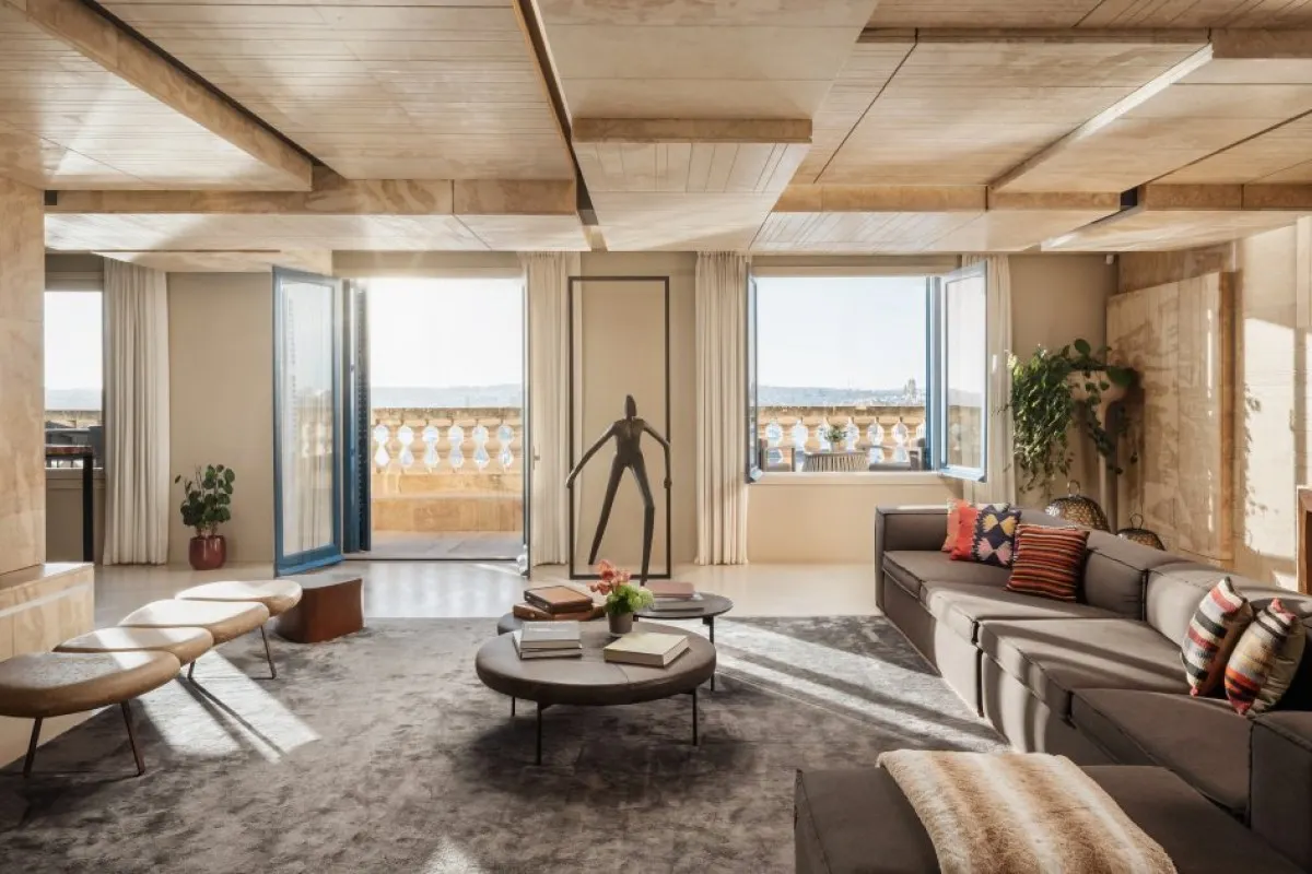 Spacious suite living room with sofa, round tables, sculpture, and open doors to a balcony overlooking Valletta rooftops