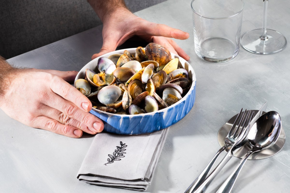 Hands placing a blue and white bowl of steamed clams on a restaurant table with cutlery and napkin