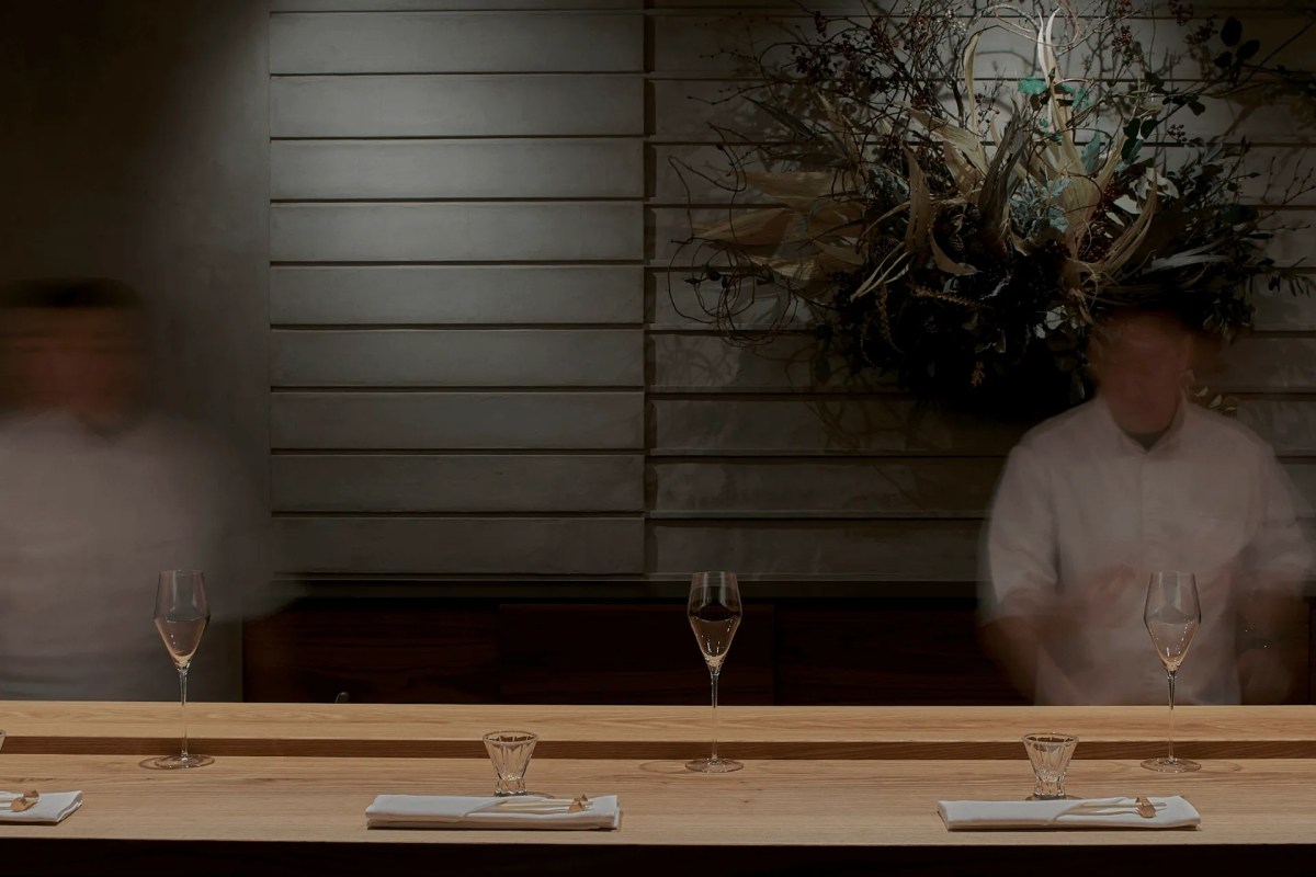 Wooden sushi counter with wine glasses and napkins, blurred chefs in background, floral wall art