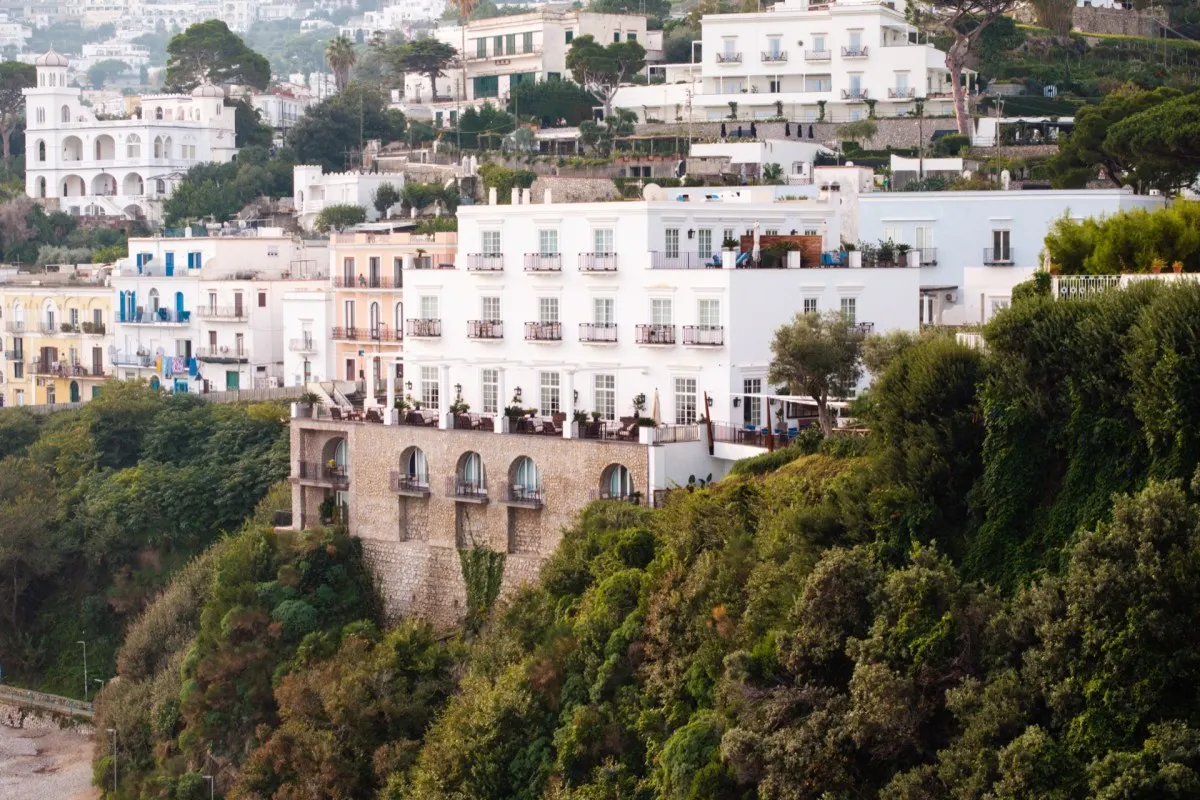 Exterior view of J.K. Place Capri's white façade perched on a cliff above the sea, surrounded by lush greenery and Capri town.