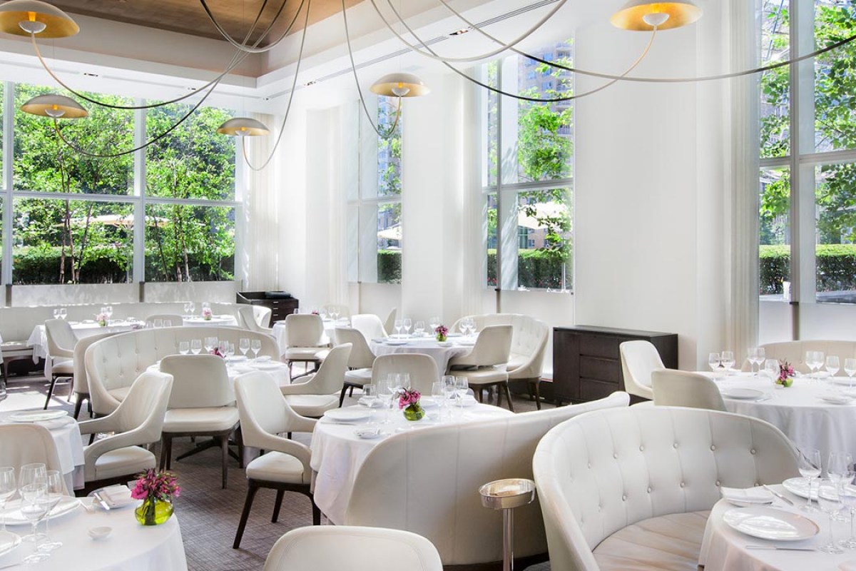 Elegant dining room with cream banquettes, round tables, and tall windows showing greenery outside.