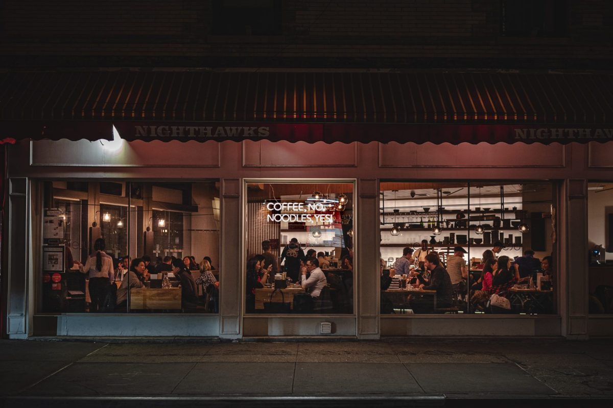 Night exterior of Jeju Noodle Bar storefront with neon sign 'Coffee, No. Noodles, Yes!' and diners visible through windows