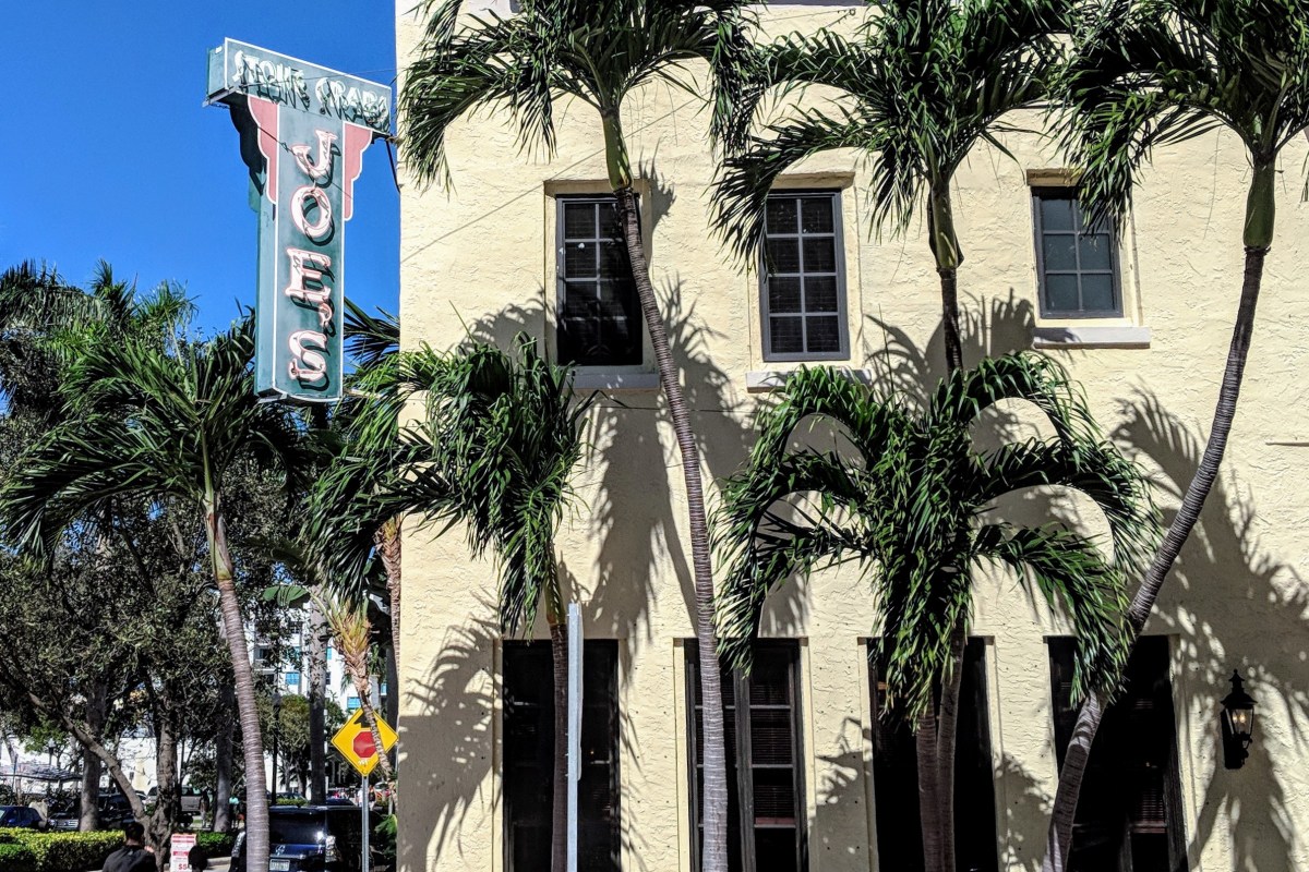 Exterior of Joe's Stone Crab with palm trees and the iconic vertical 'JOE'S' neon sign against a blue sky.