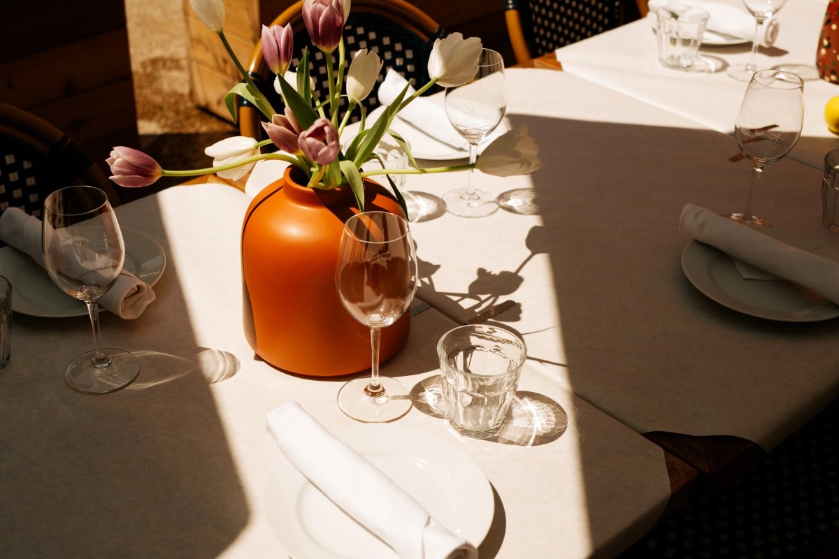 Sunlit restaurant table set with white linens, wine glasses, and an orange vase of tulips