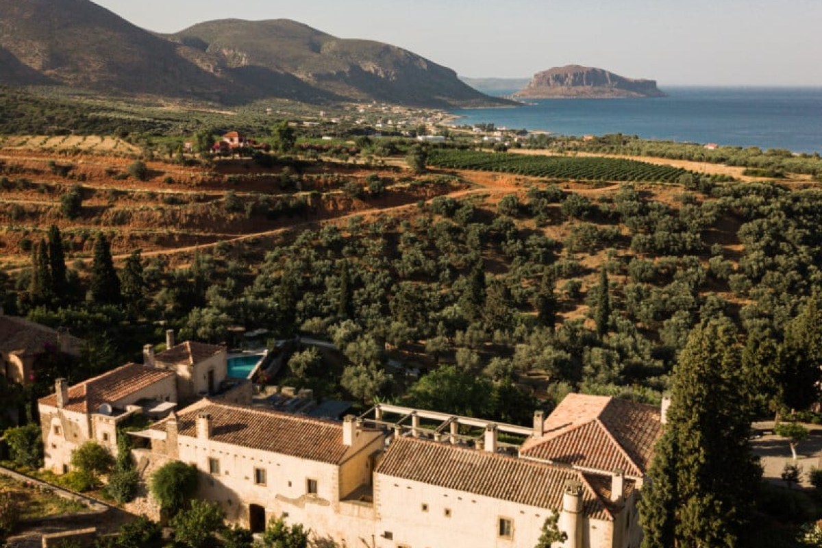 Aerial view of Kinsterna Hotel's stone buildings with sea, island, and vineyards in the background