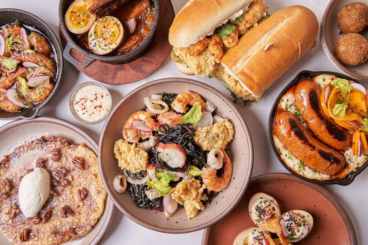 Overhead shot of various Kjun dishes including po'boy sandwiches, black noodle seafood bowl, sausages, and sides on a table