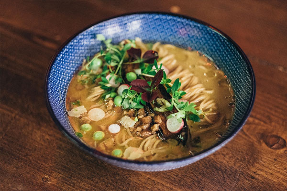 Close-up of a ramen bowl with noodles, broth, pork, peas, radish slices, and microgreens on a wooden table