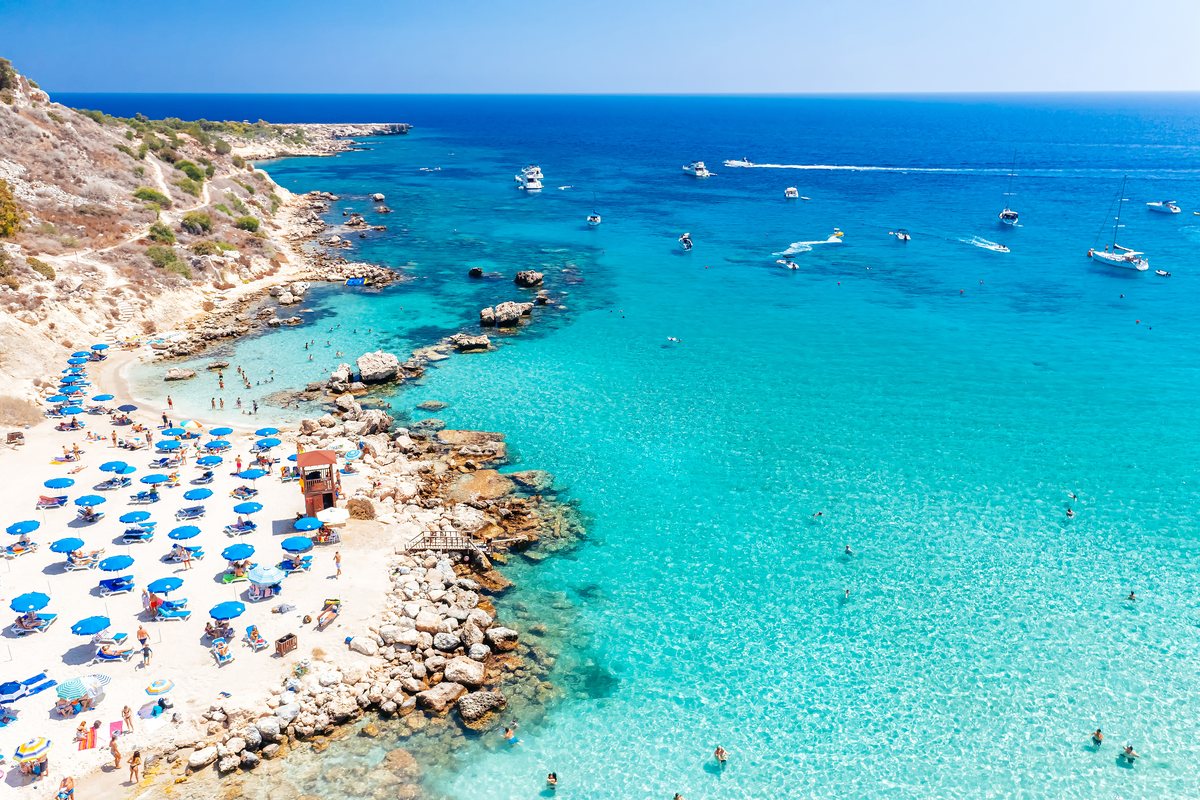 Konnos Bay near Protaras from above: a sheltered cove of turquoise water below pine-covered cliffs, with a sand beach and sunbeds