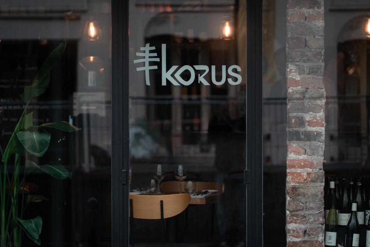 Glass entrance door of Korus restaurant with etched logo, view into dining room with set table, brick wall beside.