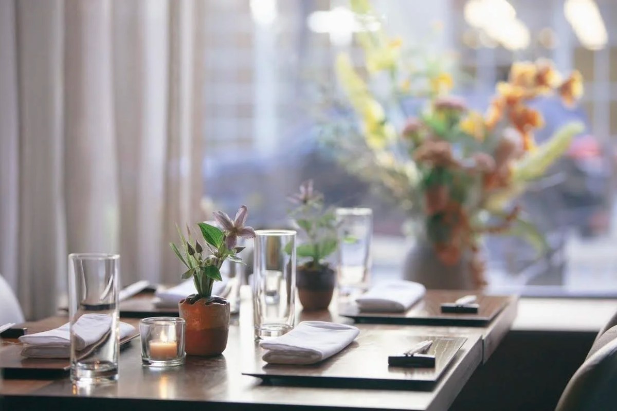 Restaurant dining table set with glasses, small potted plants, and a candle near a window with sheer curtains
