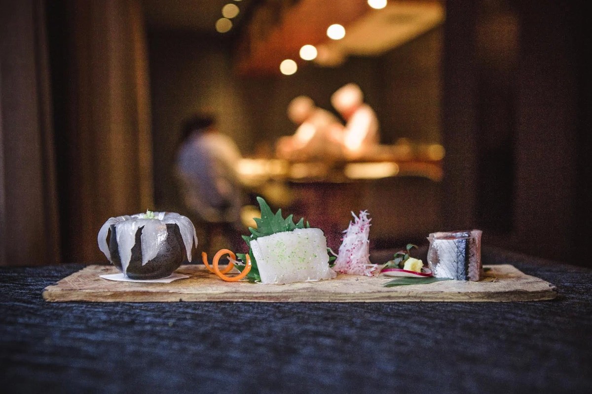 A wooden board with assorted sashimi and garnishes, with blurred chefs working at a counter behind