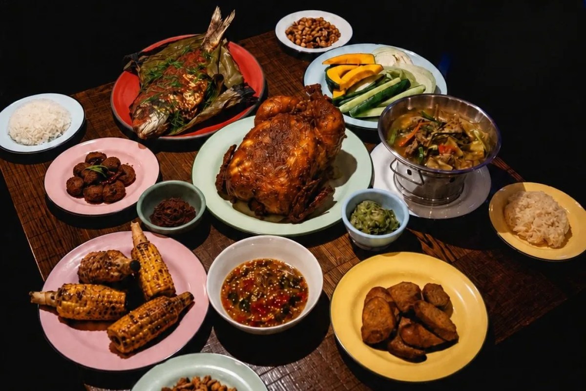 Overhead view of a Thai-style feast: whole grilled fish, roast chicken, corn, rice, curry, and sides on a dark table