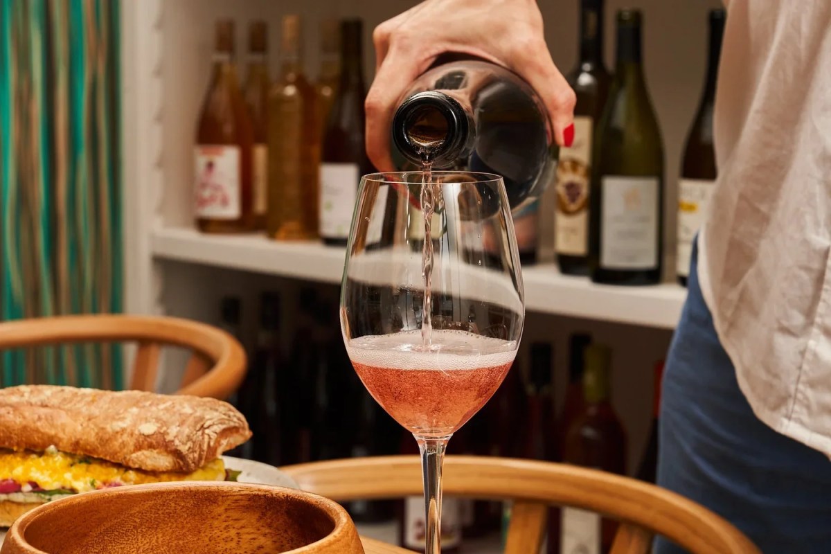 Hand pouring rosé wine into a glass at a bar area with wine bottles on shelves