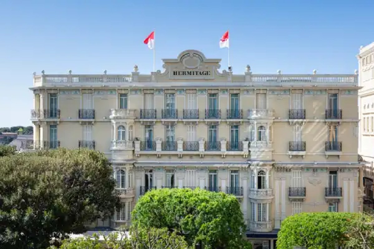 Exterior facade of the Hôtel Hermitage Monte-Carlo, cream Belle Époque building with 'HERMITAGE' sign and Monaco flags, framed by green trees