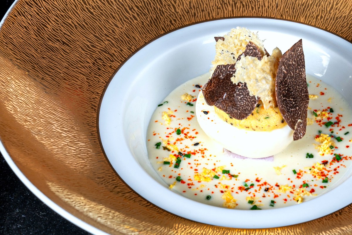 Close-up of deviled egg topped with shaved black truffle in a creamy sauce, elegant gold-rimmed bowl