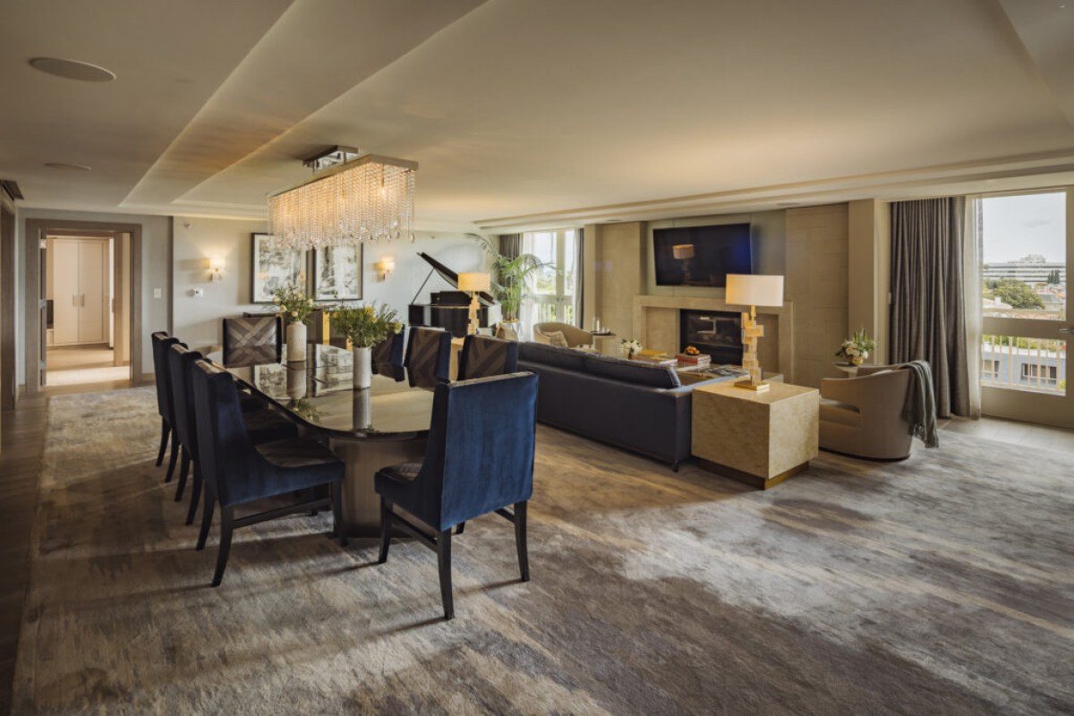 Elegant hotel suite living and dining area with piano, fireplace, and city view through open doors.
